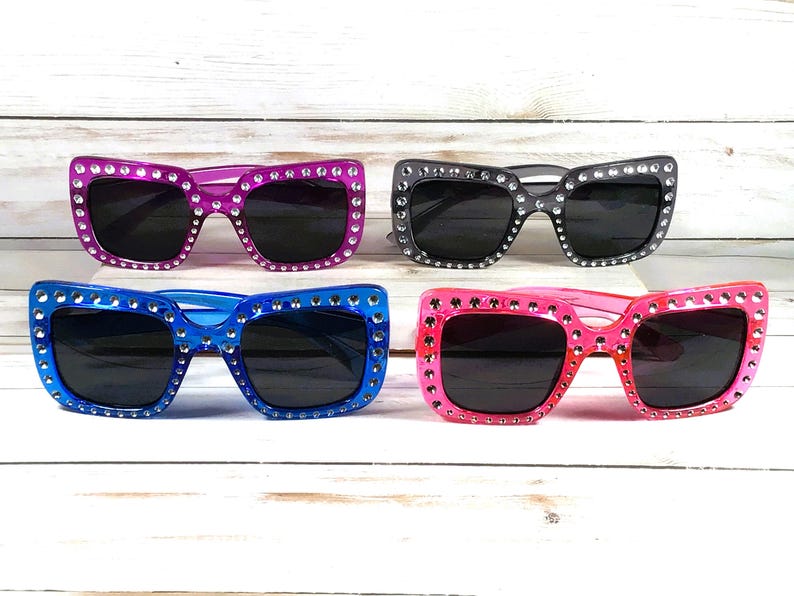 May include: Four pairs of sunglasses with rhinestones around the frames. The sunglasses are in four different colors: pink, blue, black, and purple.