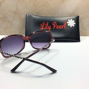 May include: Burgundy-colored sunglasses with a silver chain detail on the frame. The sunglasses are resting on a black case with the text "Lily Pearl" and a white daisy.
