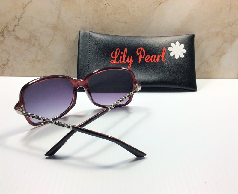 May include: Burgundy-colored sunglasses with a silver chain detail on the frame. The sunglasses are resting on a black case with the text "Lily Pearl" and a white daisy.