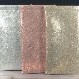 Op de afbeelding: Drie rechthoekige zakjes in zilver, ros&eacute;goud en goud, elk met een getextureerd, glinsterend oppervlak. De zakjes staan rechtop, naast elkaar, tegen een donkere achtergrond. De zakjes lijken gemaakt van een zacht, flexibel materiaal.