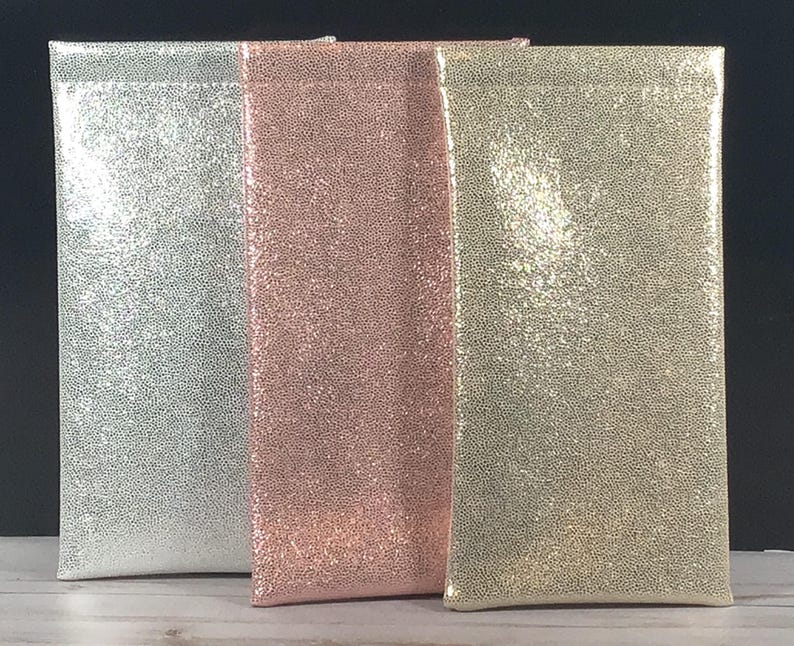 Op de afbeelding: Drie rechthoekige zakjes in zilver, ros&eacute;goud en goud, elk met een getextureerd, glinsterend oppervlak. De zakjes staan rechtop, naast elkaar, tegen een donkere achtergrond. De zakjes lijken gemaakt van een zacht, flexibel materiaal.