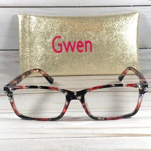 Pu&ograve; includere: Occhiali rettangolari con montatura fantasia rossa, nera e bianca e lenti trasparenti. La montatura ha strass decorativi. Dietro gli occhiali c'&egrave; un astuccio dorato con il nome "Gwen" in lettere rosa.
