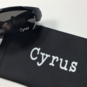 K&ouml;nnte beinhalten: Schwarze Sonnenbrille mit dunkelgrauen Gl&auml;sern und dem Wort "Cyrus" auf der Seite bedruckt. Die Sonnenbrille liegt auf einem schwarzen Etui mit dem Wort "Cyrus" in Wei&szlig; gedruckt.
