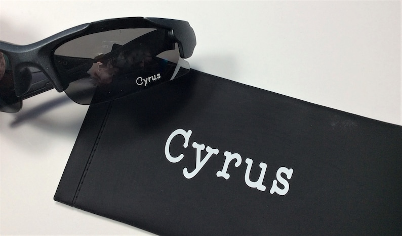K&ouml;nnte beinhalten: Schwarze Sonnenbrille mit dunkelgrauen Gl&auml;sern und dem Wort "Cyrus" auf der Seite bedruckt. Die Sonnenbrille liegt auf einem schwarzen Etui mit dem Wort "Cyrus" in Wei&szlig; gedruckt.