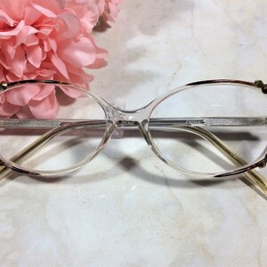 Puede incluir: Gafas transparentes con un borde y adornos decorativos dorados. Las monturas tienen una forma sutil de ojo de gato. Las patillas son de color amarillo claro. El texto "CHINA" es visible en la montura.