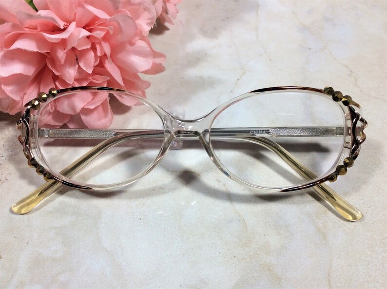 Puede incluir: Gafas transparentes con un borde y adornos decorativos dorados. Las monturas tienen una forma sutil de ojo de gato. Las patillas son de color amarillo claro. El texto "CHINA" es visible en la montura.