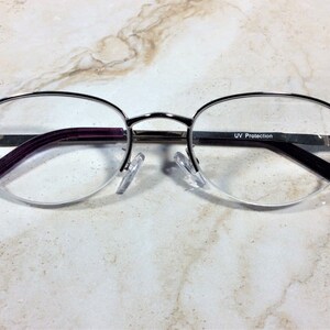May include: Silver-rimmed spectacles with a half-frame design. The arms are a deep purple, and the lenses are clear. The frame is decorated with iridescent rhinestones at the temples. The text "UV Protection" is visible on the arm. Made in China.