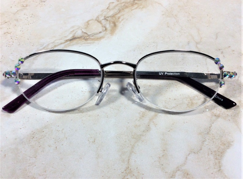 May include: Silver-rimmed spectacles with a half-frame design. The arms are a deep purple, and the lenses are clear. The frame is decorated with iridescent rhinestones at the temples. The text "UV Protection" is visible on the arm. Made in China.