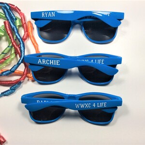 Puede incluir: Se muestran tres pares de gafas de sol azul brillante con lentes negras. Cada par tiene un nombre impreso en la montura: "RYAN", "ARCHIE" y "DABIE". El texto "WWXC 4 LIFE" tambi&eacute;n est&aacute; impreso en las monturas. Serpentinas de colores en el fondo.
