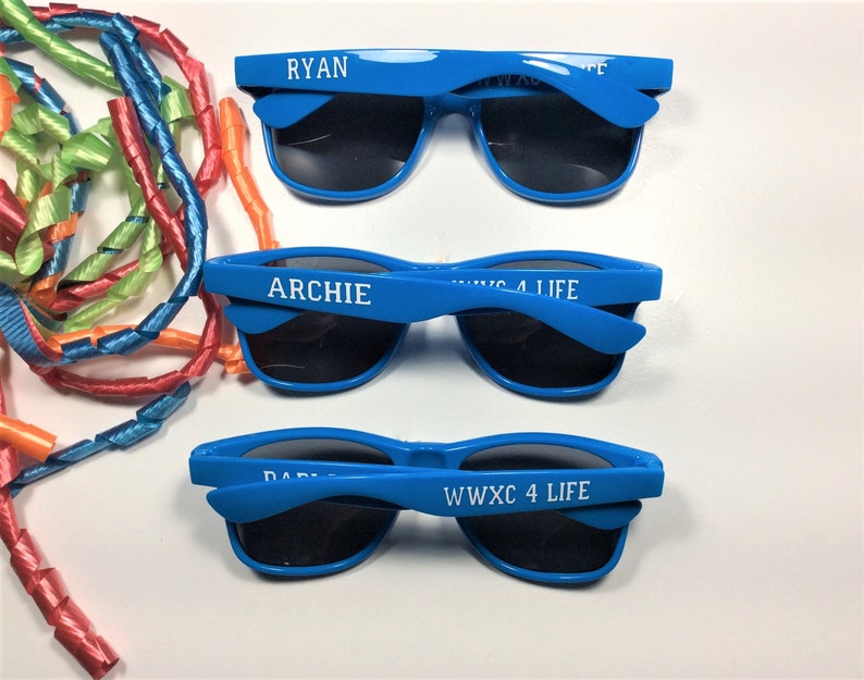 Puede incluir: Se muestran tres pares de gafas de sol azul brillante con lentes negras. Cada par tiene un nombre impreso en la montura: "RYAN", "ARCHIE" y "DABIE". El texto "WWXC 4 LIFE" tambi&eacute;n est&aacute; impreso en las monturas. Serpentinas de colores en el fondo.