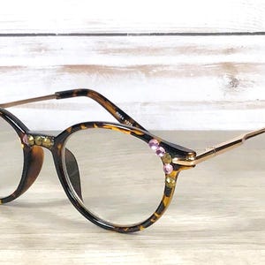 May include: Oval-shaped eyeglasses with a tortoiseshell pattern frame, adorned with gold and pink rhinestones. The glasses have gold-colored arms and black earpieces, resting on a light wooden surface.