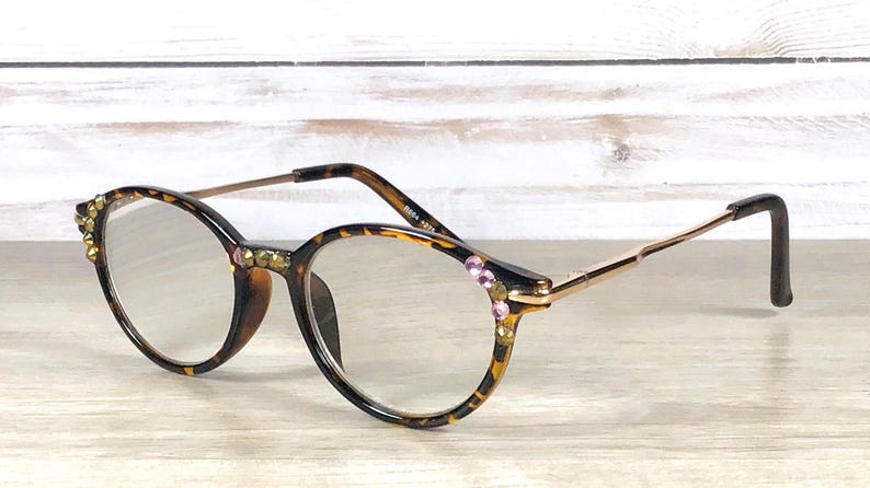 May include: Oval-shaped eyeglasses with a tortoiseshell pattern frame, adorned with gold and pink rhinestones. The glasses have gold-colored arms and black earpieces, resting on a light wooden surface.