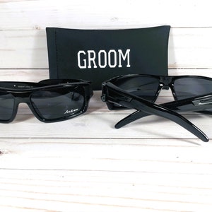 May include: Two pairs of black sunglasses with dark lenses, one with the name "Andrew" printed on the lens. A black case with the word "GROOM" in white is behind the sunglasses. The sunglasses are on a light wood surface.