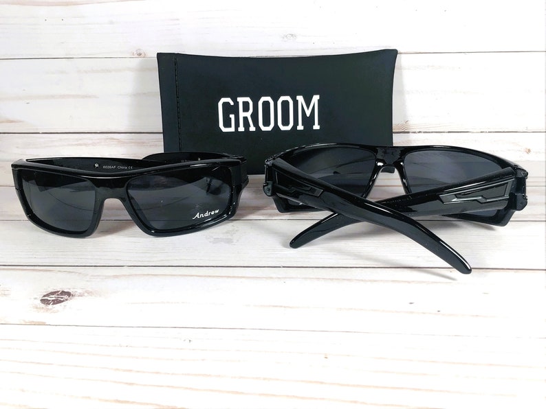 May include: Two pairs of black sunglasses with dark lenses, one with the name "Andrew" printed on the lens. A black case with the word "GROOM" in white is behind the sunglasses. The sunglasses are on a light wood surface.