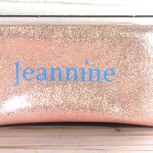 Pu&ograve; includere: Una pochette rettangolare color oro rosa con una superficie strutturata e maculata. Il nome "Jeannine" &egrave; ricamato in blu chiaro al centro. La pochette &egrave; probabilmente realizzata in un materiale morbido e flessibile.