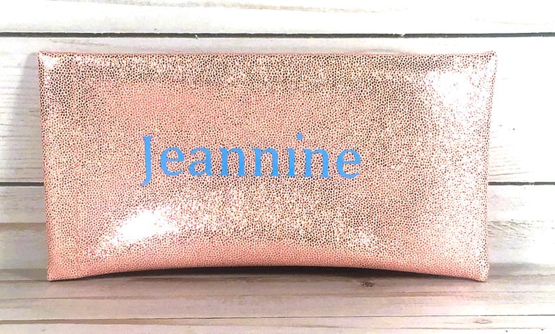 Pu&ograve; includere: Una pochette rettangolare color oro rosa con una superficie strutturata e maculata. Il nome "Jeannine" &egrave; ricamato in blu chiaro al centro. La pochette &egrave; probabilmente realizzata in un materiale morbido e flessibile.