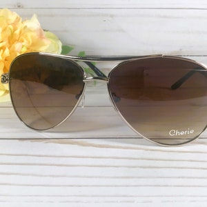 Peut inclure: Lunettes de soleil aviateur dor&eacute;es avec des verres d&eacute;grad&eacute;s bruns. Les lunettes ont une monture en m&eacute;tal argent&eacute; et le mot "Cherie" est imprim&eacute; sur la lentille droite.