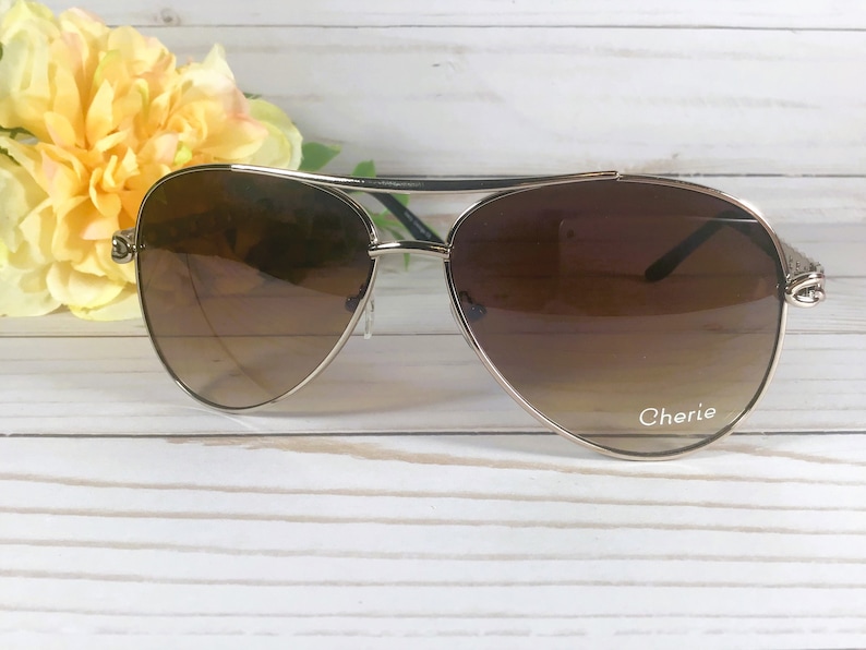 Peut inclure: Lunettes de soleil aviateur dor&eacute;es avec des verres d&eacute;grad&eacute;s bruns. Les lunettes ont une monture en m&eacute;tal argent&eacute; et le mot "Cherie" est imprim&eacute; sur la lentille droite.