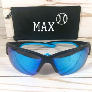 May include: Black sports sunglasses with reflective blue lenses and bright blue inner temple accents. Behind the sunglasses sits a black drawstring pouch featuring the white text MAX and a white graphic illustration of a baseball.