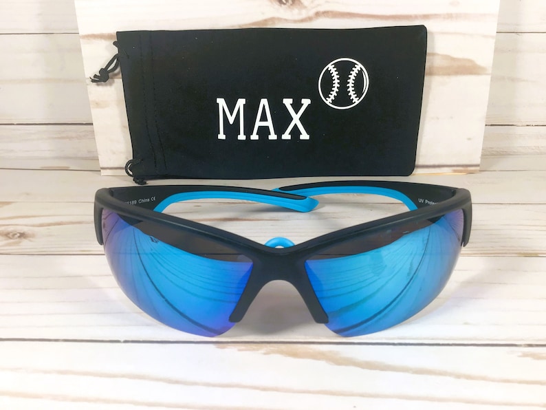 May include: Black sports sunglasses with reflective blue lenses and bright blue inner temple accents. Behind the sunglasses sits a black drawstring pouch featuring the white text MAX and a white graphic illustration of a baseball.
