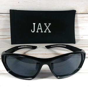 May include: Black sunglasses with a curved frame and dark lenses, resting in front of a black case. The case has the word "JAX" printed in silver. The sunglasses have a sporty design.