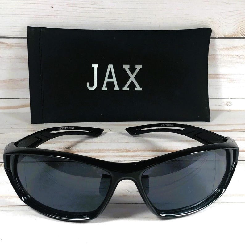 May include: Black sunglasses with a curved frame and dark lenses, resting in front of a black case. The case has the word "JAX" printed in silver. The sunglasses have a sporty design.