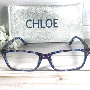 Peut inclure: Lunettes rectangulaires avec une monture &agrave; motif &eacute;caille de tortue bleu et violet. Derri&egrave;re les lunettes se trouve un &eacute;tui rectangulaire argent&eacute; &agrave; paillettes avec le mot CHLOE imprim&eacute; en lettres bleu fonc&eacute; sur la face avant de l'&eacute;tui.