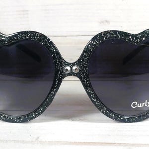 May include: Black heart-shaped sunglasses with rhinestones and a gold crown detail. The sunglasses have a sparkly finish and the text "Curly" is printed on the right lens.