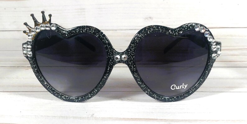 May include: Black heart-shaped sunglasses with rhinestones and a gold crown detail. The sunglasses have a sparkly finish and the text "Curly" is printed on the right lens.