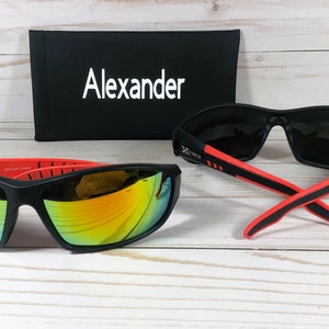 May include: Two pairs of black sunglasses with red accents. One pair has mirrored lenses and the other has dark lenses. The sunglasses are on a white wooden surface. The sunglasses come with a black case that has the word "Alexander" printed on it.