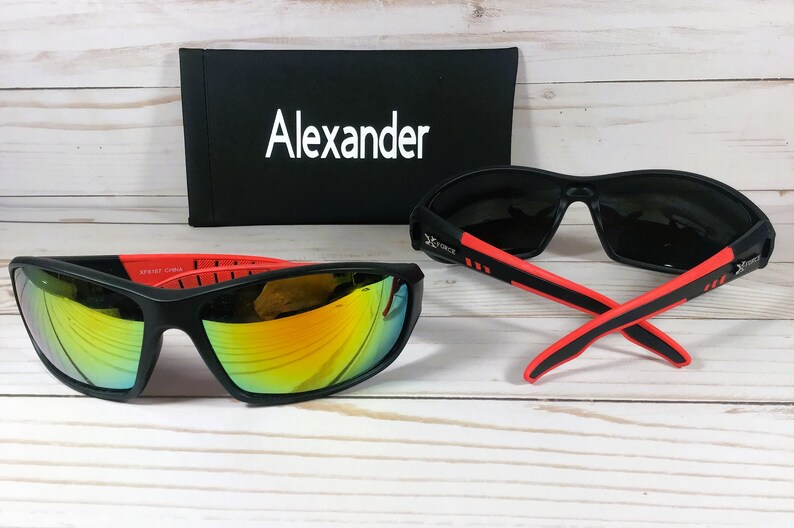 May include: Two pairs of black sunglasses with red accents. One pair has mirrored lenses and the other has dark lenses. The sunglasses are on a white wooden surface. The sunglasses come with a black case that has the word "Alexander" printed on it.