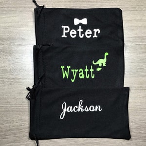 May include: Three black drawstring bags with personalized names. The top bag has a white bow tie and the name "Peter". The middle bag has a green dinosaur and the name "Wyatt". The bottom bag has the name "Jackson".
