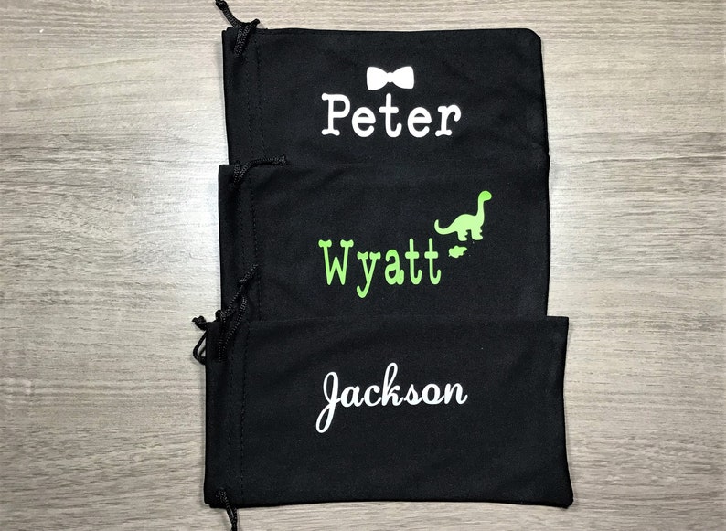 May include: Three black drawstring bags with personalized names. The top bag has a white bow tie and the name "Peter". The middle bag has a green dinosaur and the name "Wyatt". The bottom bag has the name "Jackson".