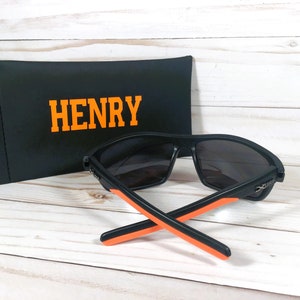 Peut inclure: Lunettes de soleil noires avec des d&eacute;tails orange et un &eacute;tui noir. L'&eacute;tui porte le nom "HENRY" en lettres orange. Les lunettes de soleil ont un design sportif.