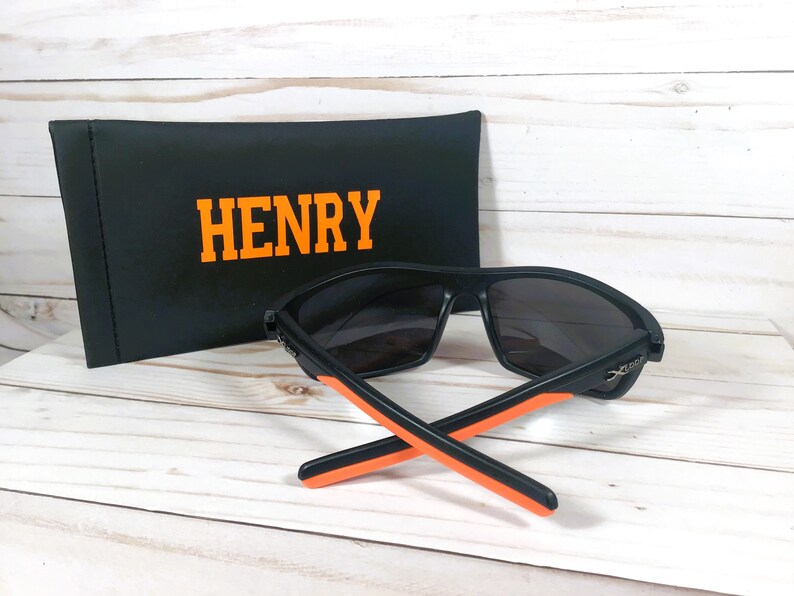 Peut inclure: Lunettes de soleil noires avec des d&eacute;tails orange et un &eacute;tui noir. L'&eacute;tui porte le nom "HENRY" en lettres orange. Les lunettes de soleil ont un design sportif.