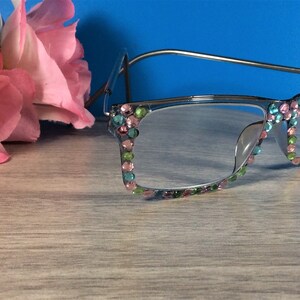 May include: Silver-framed reading glasses with colorful rhinestones. The rhinestones are in shades of pink, green, and blue, adorning the frame's edges. A pink flower is in the background. The glasses are on a light wood-grain surface.