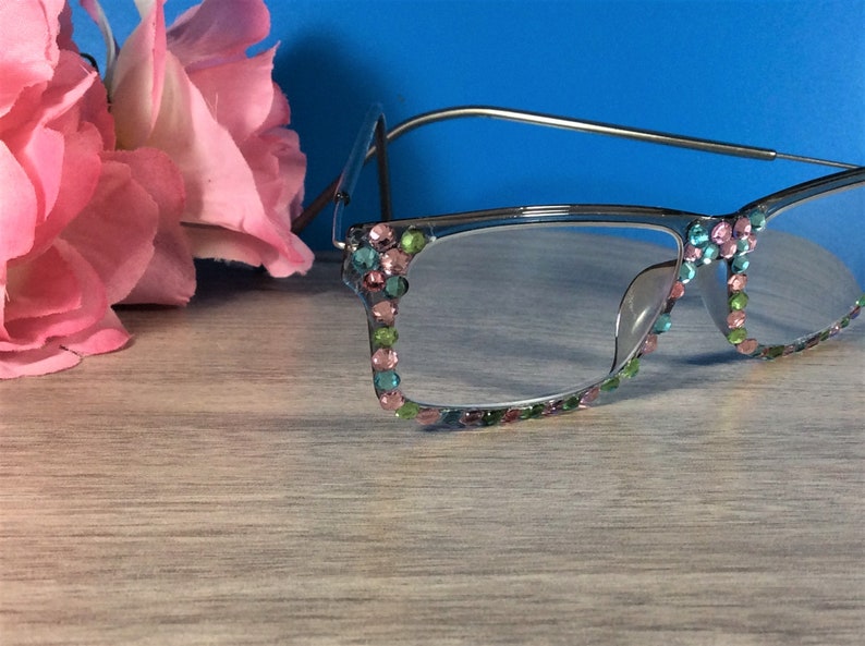 May include: Silver-framed reading glasses with colorful rhinestones. The rhinestones are in shades of pink, green, and blue, adorning the frame's edges. A pink flower is in the background. The glasses are on a light wood-grain surface.