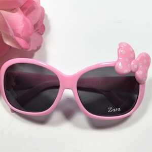 Puede incluir: Gafas de sol de pl&aacute;stico rosa con un lazo blanco de lunares en el lado derecho. Las gafas tienen lentes oscuros y dicen "Zara" en la lente derecha.