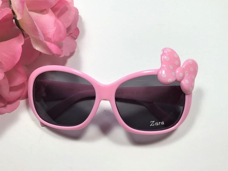 Puede incluir: Gafas de sol de pl&aacute;stico rosa con un lazo blanco de lunares en el lado derecho. Las gafas tienen lentes oscuros y dicen "Zara" en la lente derecha.