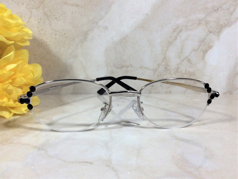 May include: Silver-rimmed eyeglasses with a half-frame design. The frames feature black embellishments on the outer corners. The lenses are clear, and the bridge and temples are a silver and black color. The text "2319 +1.50" is printed on the frame.