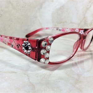 Peut inclure: Lunettes &agrave; monture rose avec verres transparents. Les montures sont orn&eacute;es de strass transparents et d'un motif floral sur les branches. Une gemme noire est centr&eacute;e dans une fleur sur la monture.