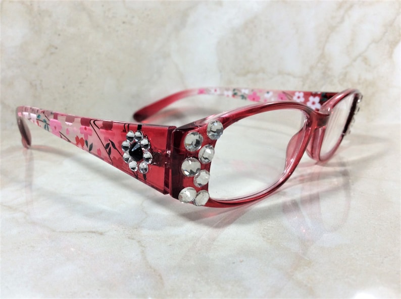 Peut inclure: Lunettes &agrave; monture rose avec verres transparents. Les montures sont orn&eacute;es de strass transparents et d'un motif floral sur les branches. Une gemme noire est centr&eacute;e dans une fleur sur la monture.