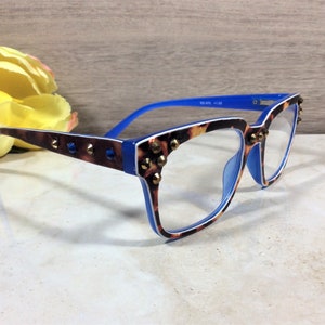 May include: Eyeglasses with a blue and tortoiseshell pattern frame, accented with gold-coloured studs. The arms are blue, and the lenses are clear. The glasses are set against a neutral background with a yellow flower.