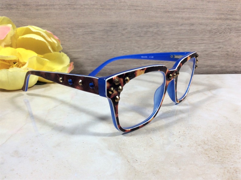 May include: Eyeglasses with a blue and tortoiseshell pattern frame, accented with gold-coloured studs. The arms are blue, and the lenses are clear. The glasses are set against a neutral background with a yellow flower.