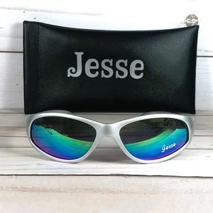 Op de afbeelding: Zilverkleurige zonnebril met iriserende, meerkleurig reflecterende glazen met de naam Jesse op het rechterglas gedrukt. De zonnebril wordt geleverd met een zwart rechthoekig beschermend etui met de naam Jesse in zilverkleurige letters.