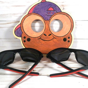 Peut inclure: Deux paires de lunettes de soleil noires avec des accents rouges sur les branches, devant une d&eacute;coupe de visage de grenouille de dessin anim&eacute;. Les lunettes de soleil ont des verres fonc&eacute;s et un design sportif. La d&eacute;coupe de grenouille est peinte en orange, marron et violet.