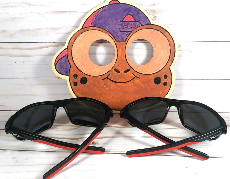 Peut inclure: Deux paires de lunettes de soleil noires avec des accents rouges sur les branches, devant une d&eacute;coupe de visage de grenouille de dessin anim&eacute;. Les lunettes de soleil ont des verres fonc&eacute;s et un design sportif. La d&eacute;coupe de grenouille est peinte en orange, marron et violet.