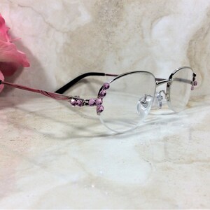 May include: Silver-rimmed spectacles with pink and black arms. The frames are decorated with pink gemstones. The clear lenses are set against a marble background, with a pink flower in the background.