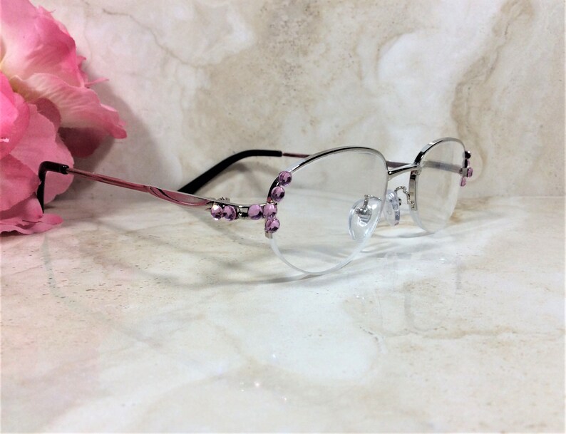 May include: Silver-rimmed spectacles with pink and black arms. The frames are decorated with pink gemstones. The clear lenses are set against a marble background, with a pink flower in the background.