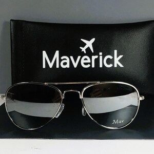 May include: Aviator sunglasses with thin silver metal frames and reflective silver lenses. The right lens features the text Mav in white. Behind the sunglasses is a black rectangular case with the word Maverick and an airplane icon.